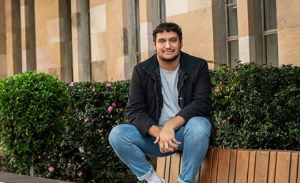 A man sits on the back of an outdoor bench with a garden and sandstone building in the background.